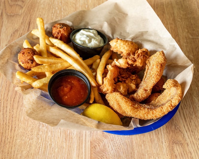Fried fish, shrimp, hush puppies, and french fries with dipping sauces and a lemon wedge.