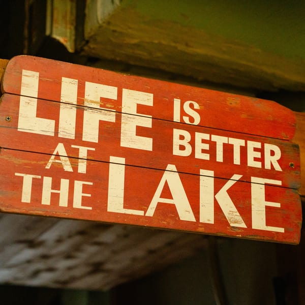 A rustic red wooden sign with white text reads, "LIFE IS BETTER AT THE LAKE".