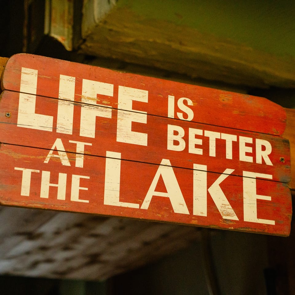 A rustic red wooden sign with white text reads, "LIFE IS BETTER AT THE LAKE".