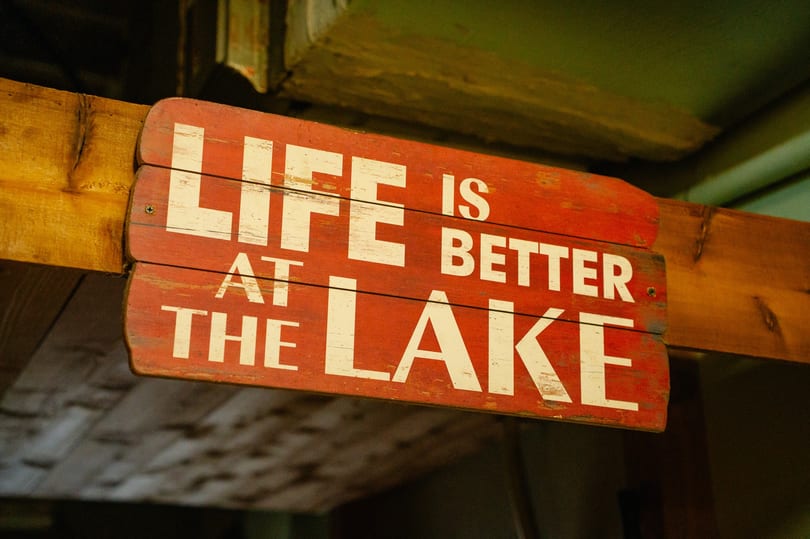 A rustic red wooden sign with white text reads, "LIFE IS BETTER AT THE LAKE".