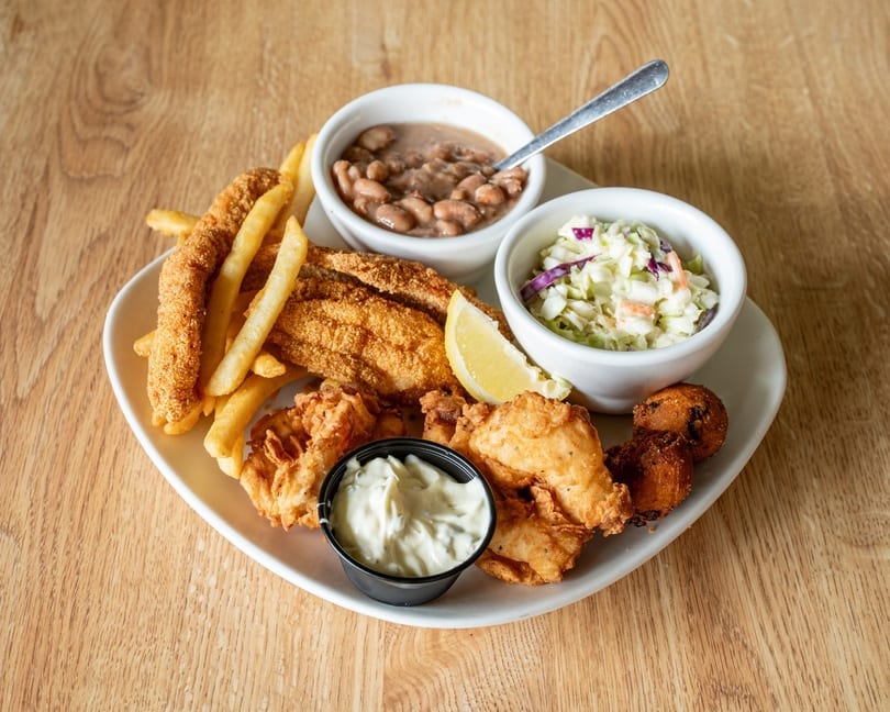 Fried fish, shrimp, fries, hushpuppies, coleslaw, baked beans, and tartar sauce on a white plate.