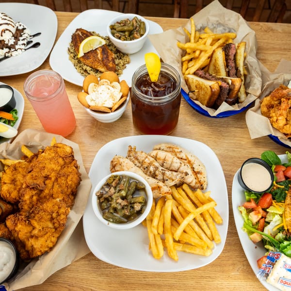 A wooden table laden with various dishes, including fried chicken, grilled fish, salads, fries, and desserts.