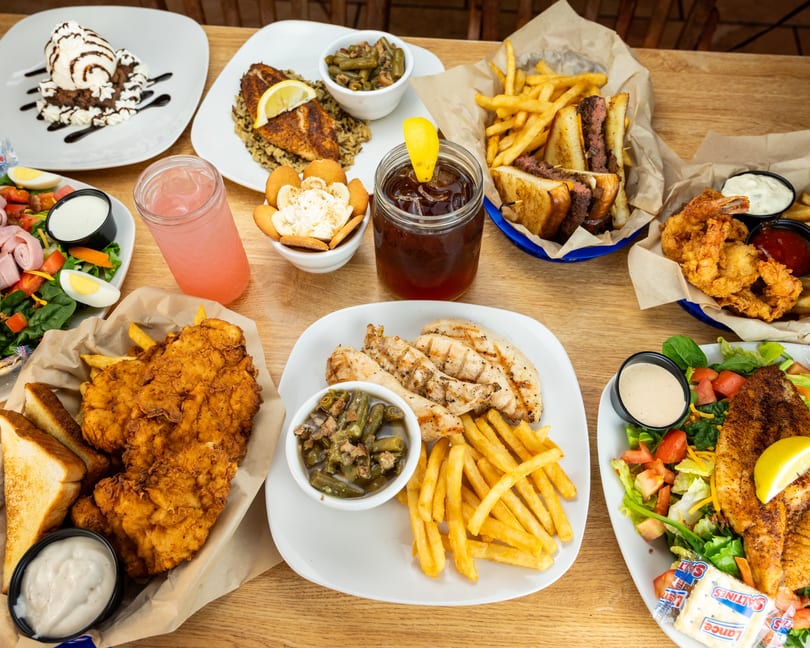 A wooden table laden with various dishes, including fried chicken, grilled fish, salads, fries, and desserts.