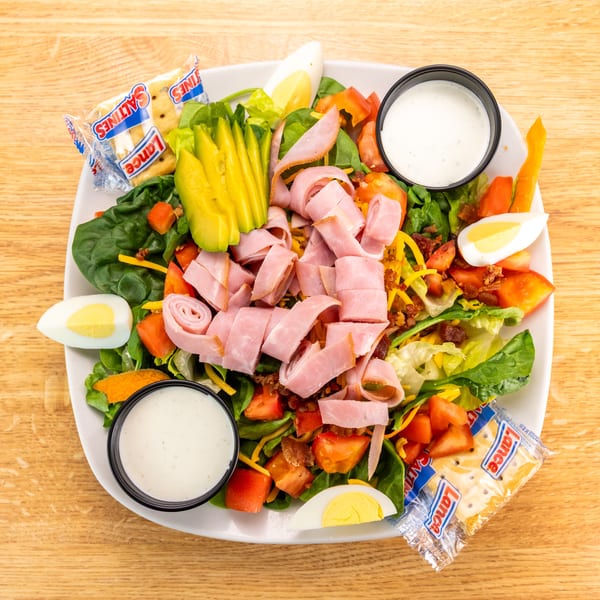 Overhead view of a chef salad with ham, avocado, eggs, tomatoes, and crackers on a wooden table.