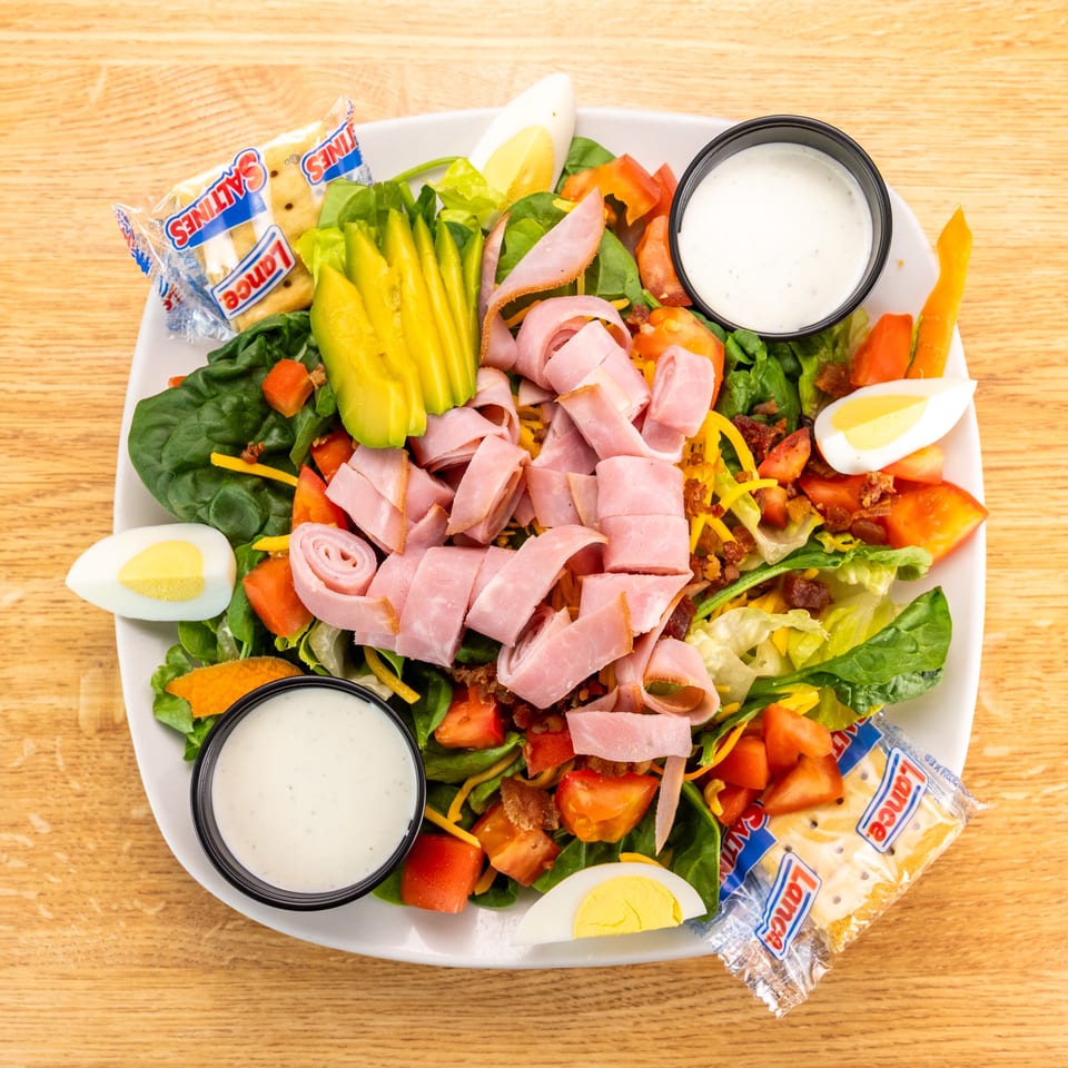 Overhead view of a chef salad with ham, avocado, eggs, tomatoes, and crackers on a wooden table.