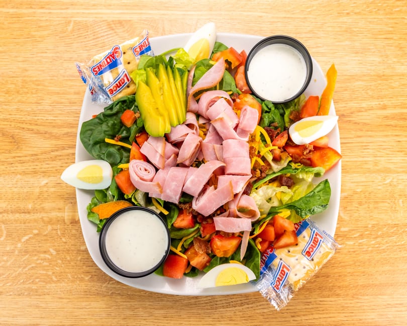 Overhead view of a chef salad with ham, avocado, eggs, tomatoes, and crackers on a wooden table.