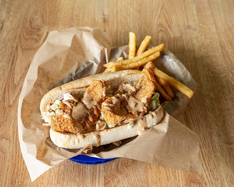 Fried shrimp po'boy sandwich with coleslaw and sauce, served with french fries on a wooden table.