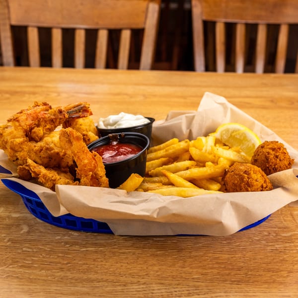 Fried shrimp, french fries, hush puppies, and dipping sauces in a blue basket on a wooden table.