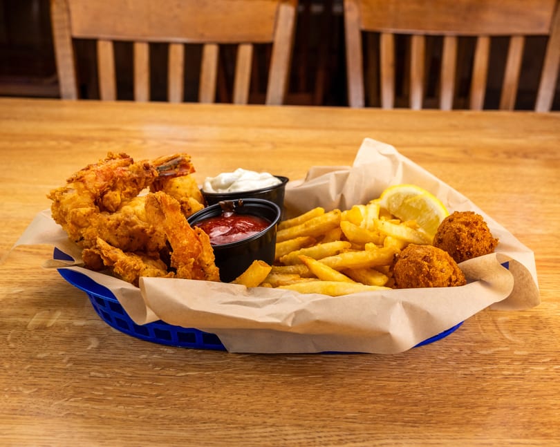 Fried shrimp, french fries, hush puppies, and dipping sauces in a blue basket on a wooden table.