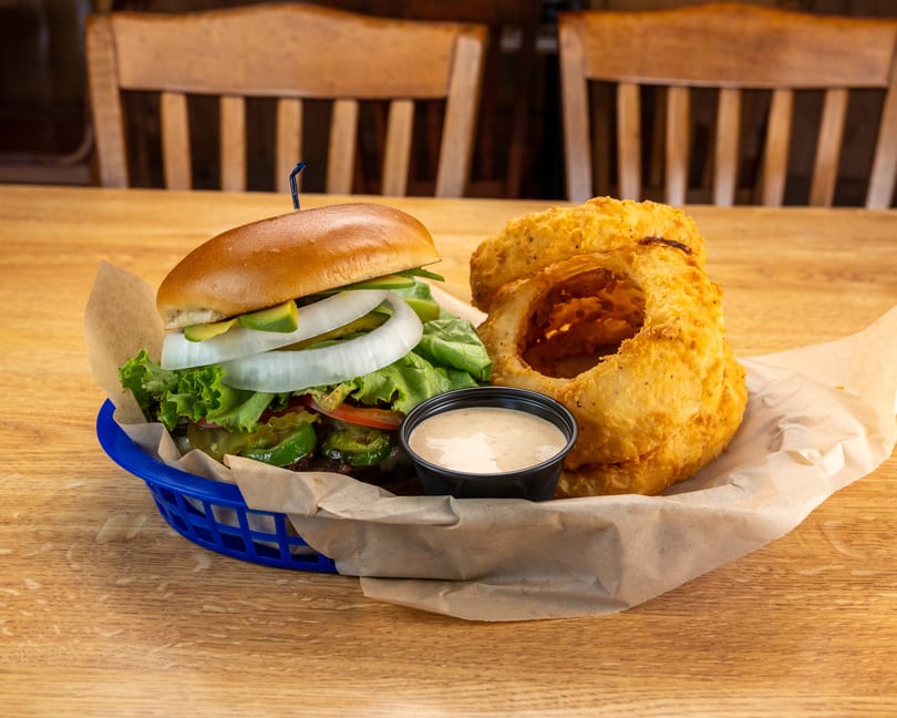 A burger with avocado, onion, lettuce, tomato, and jalapeños, served with onion rings and dipping sauce.
