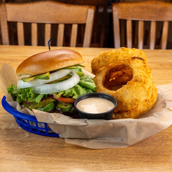 A burger with avocado, onion, lettuce, and jalapeños, served with onion rings and dipping sauce.